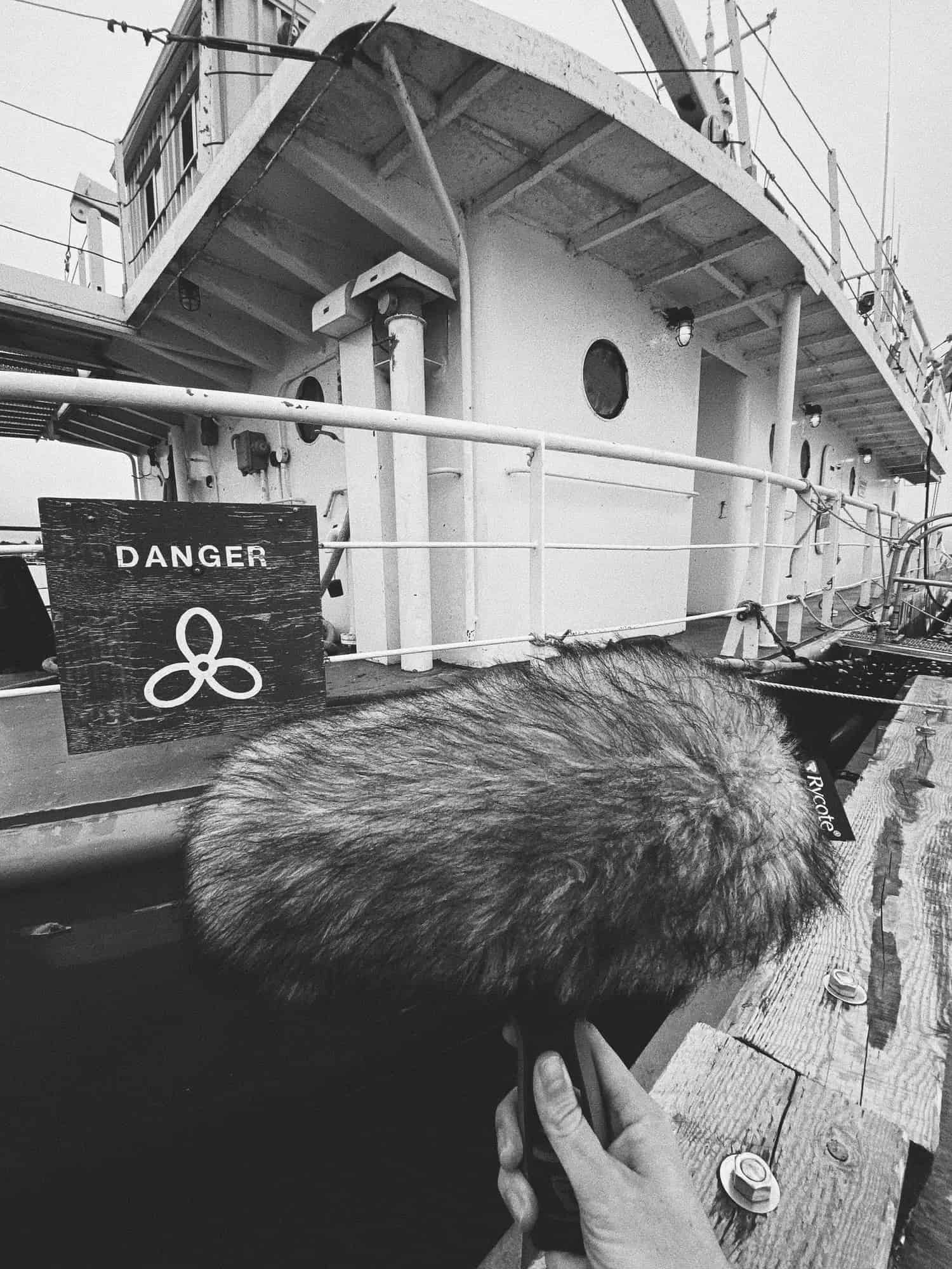 Black and white photograph of a hand holding a microphone inside a wind guard in front of a large ship.